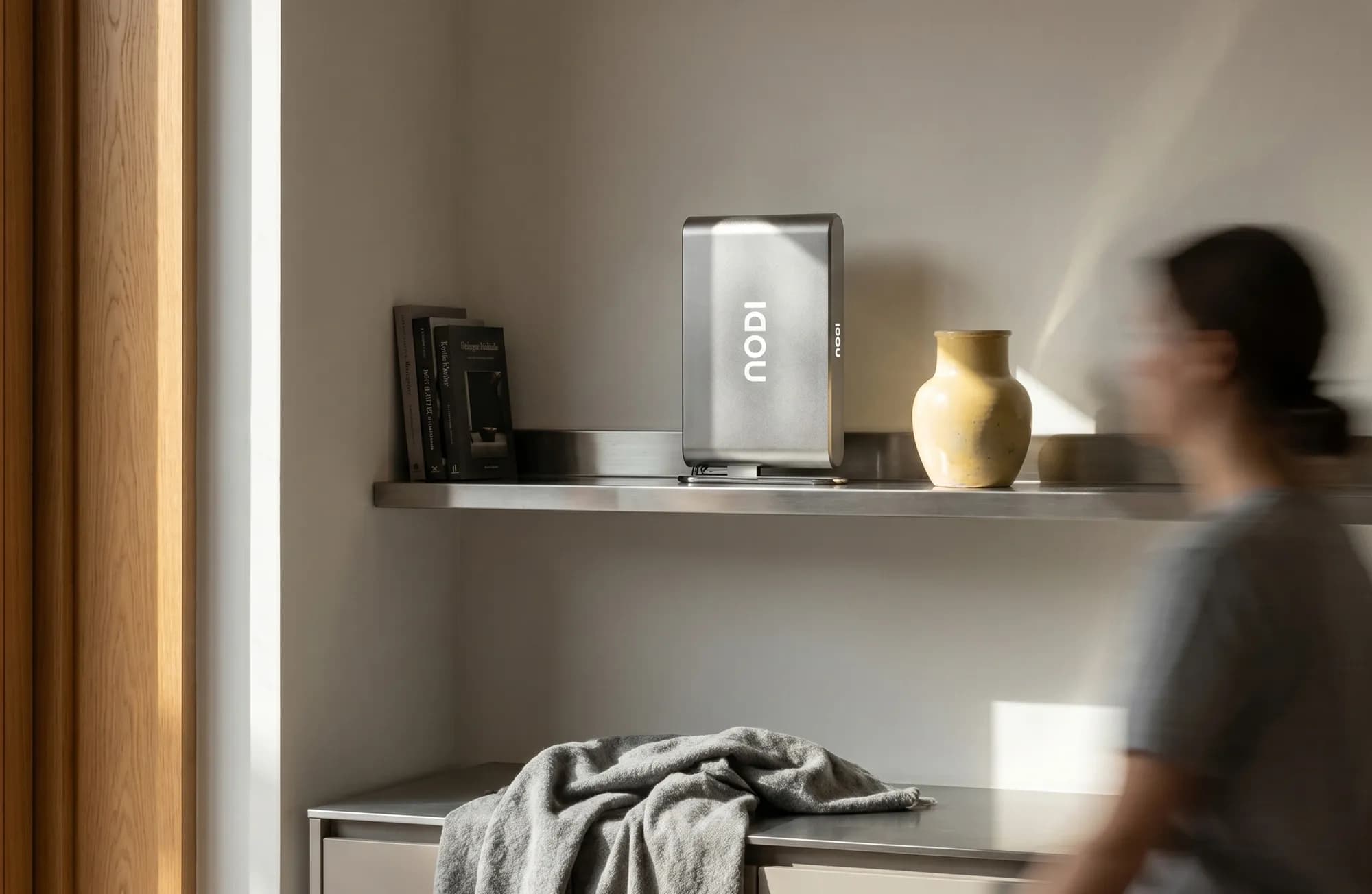 NODI battery on a stainless steel shelf in a kitchen beside a yellow ceramic vase and books. A blurred woman walks past in the foreground and a grey cloth lies on the counter below. Soft side light.