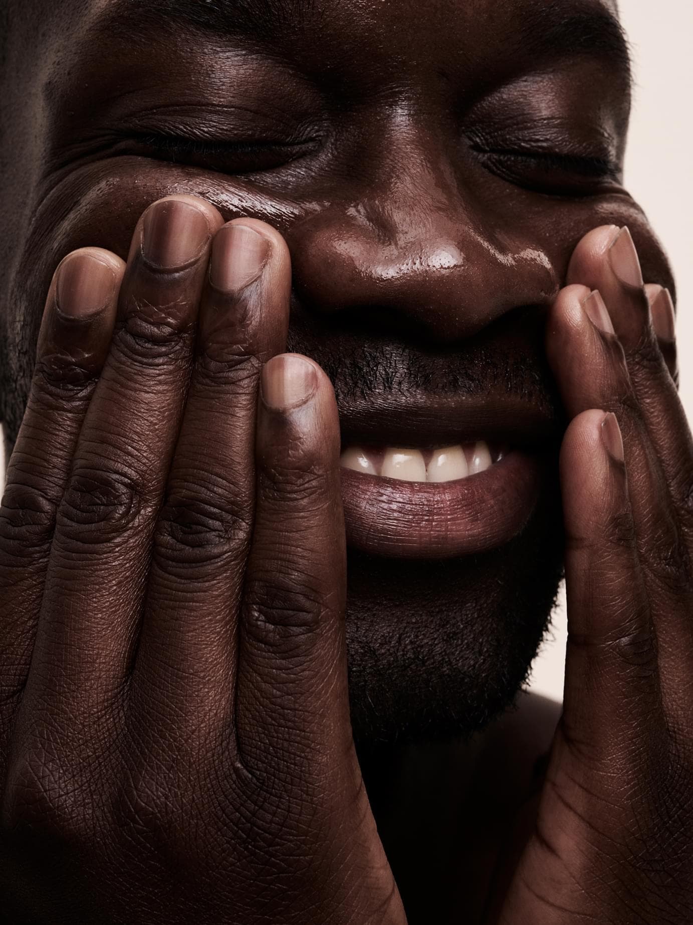 Extreme close-up of a man's face with both hands pressed to his cheeks; glossy, moisturized skin and a broad smile convey the product's caring effect.
